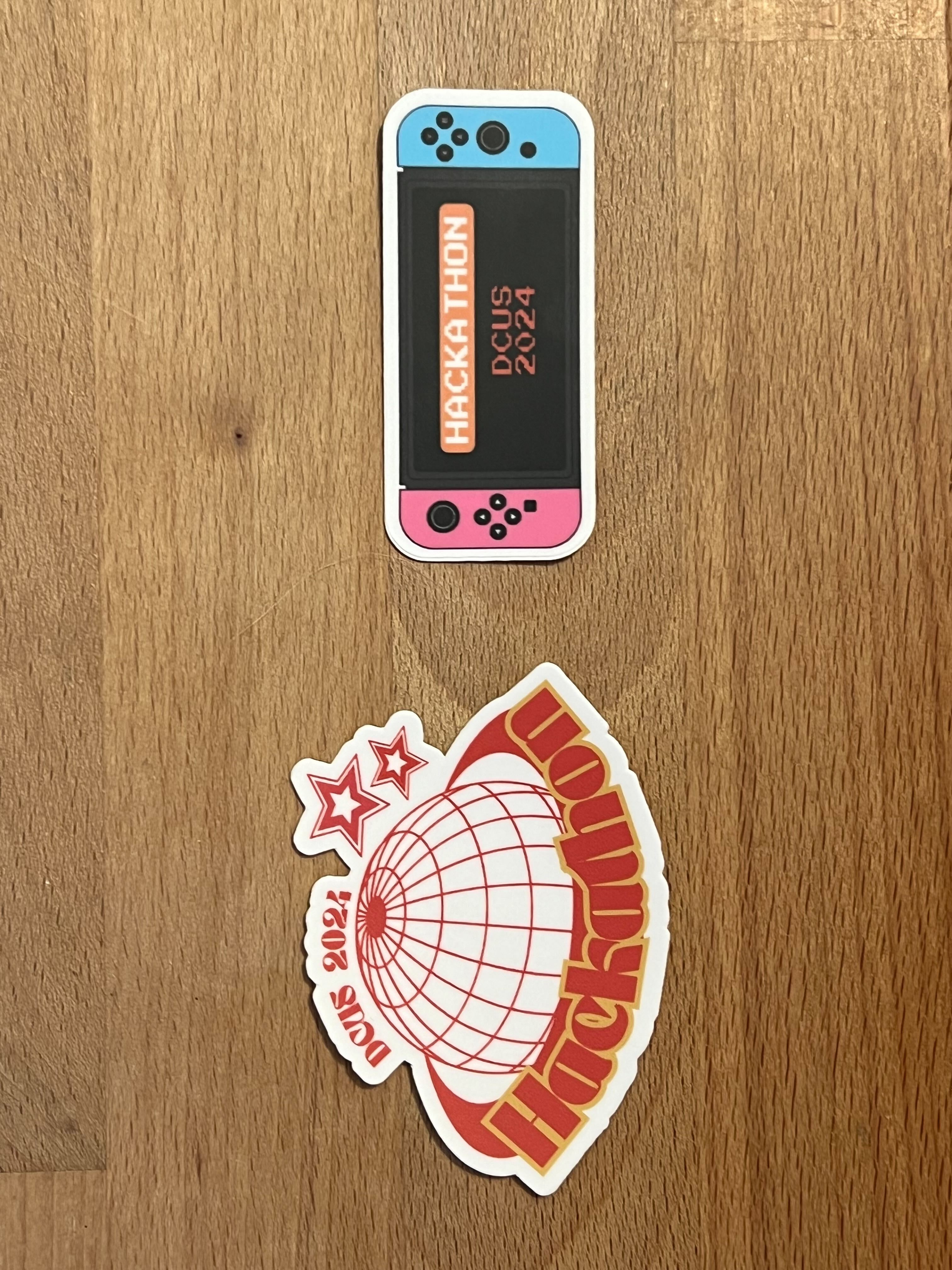 Two hackathon stickers. Both say DCUS 2024 Hackathon. The first is a red and yellow sphere with stars to the side. The second is a pink and blue game console.