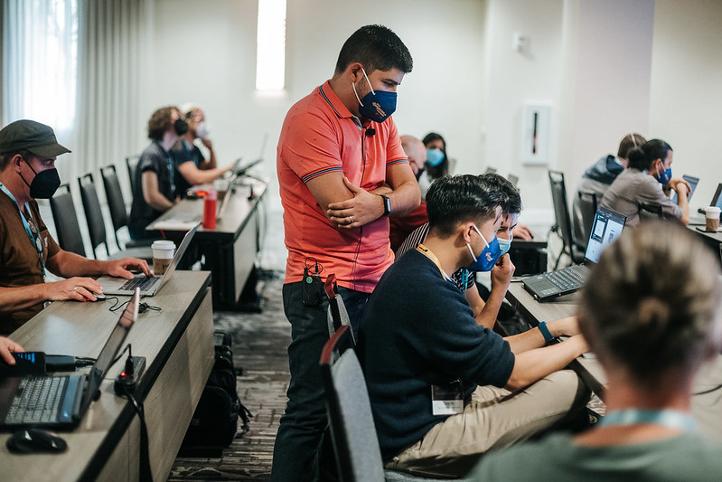 A presenter at DjangoCon US 2022 helping a pair of people working at their laptop. There are several other people in the background at their desks working on laptops.